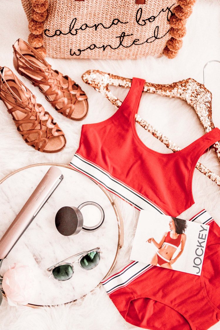 A flatlay display of festival staple sandals, flat iron, sunglasses, moisturizer, and red retro Jockey underwear with a straw bag that reads "cabana boy wanted"