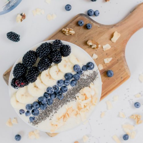 smoothie bowl with blueberries, banana slices, coconut flakes, chia seeds, and blackberries on a cutting board and white counter