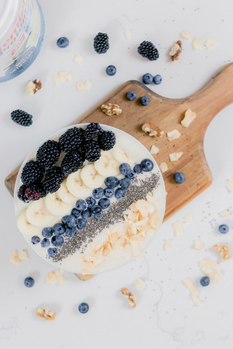 smoothie bowl with blueberries, banana slices, coconut flakes, chia seeds, and blackberries on a cutting board and white counter